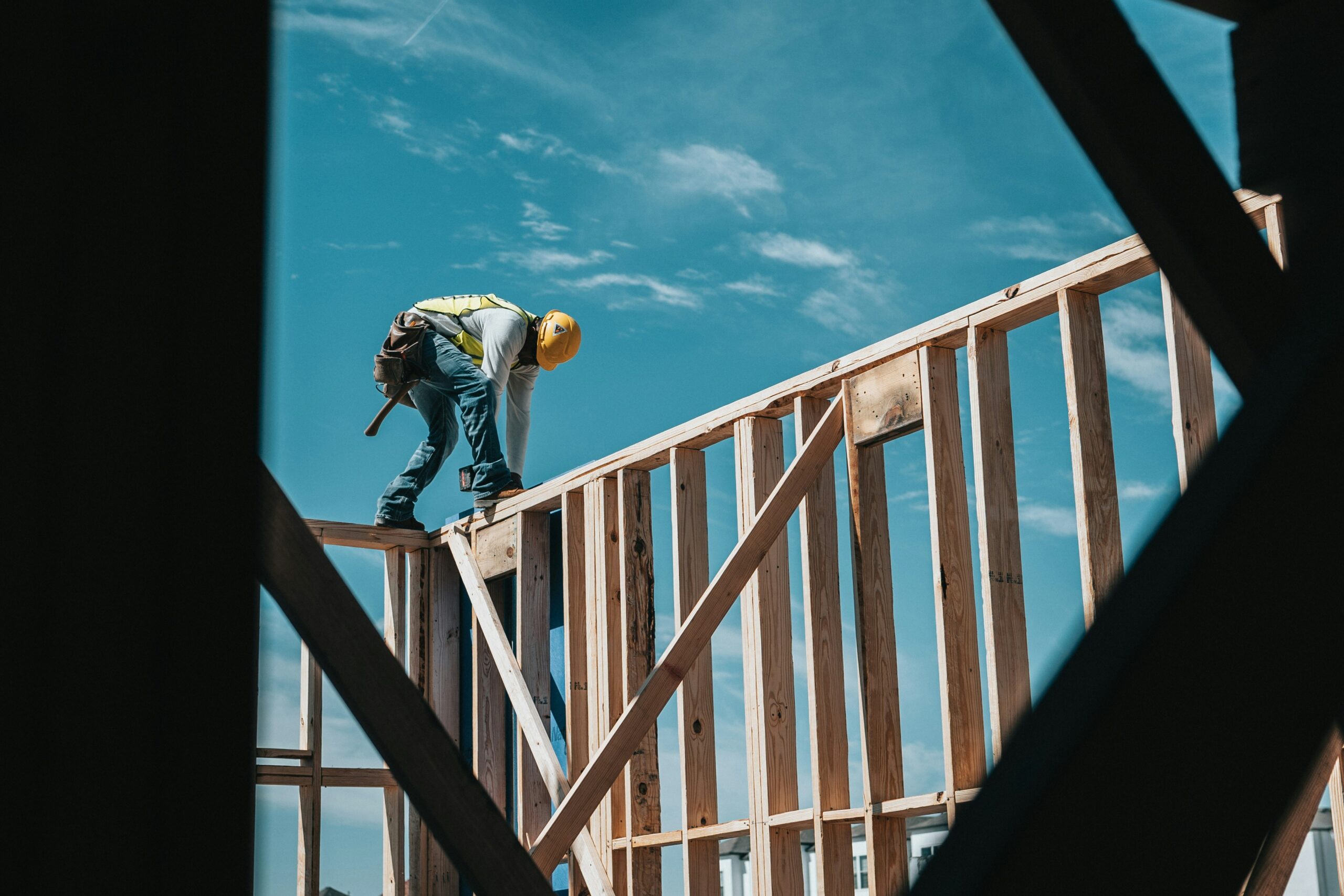 Construction worker wearing a hard hat and safety vest standing on a wooden frame of a building under construction, with a clear blue sky in the background. The image is framed by dark, angular elements in the foreground.