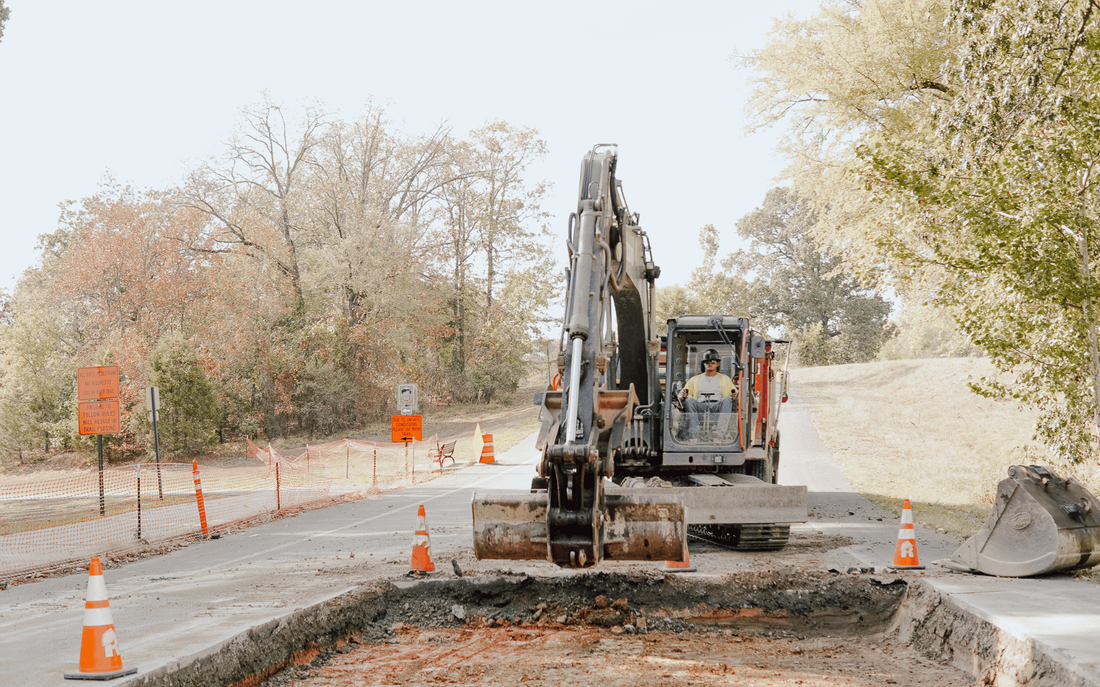 11 A construction worker operates a large excavator on a blocked-off road, surrounded by traffic cones and signs, with trees and safety fencing in the background.