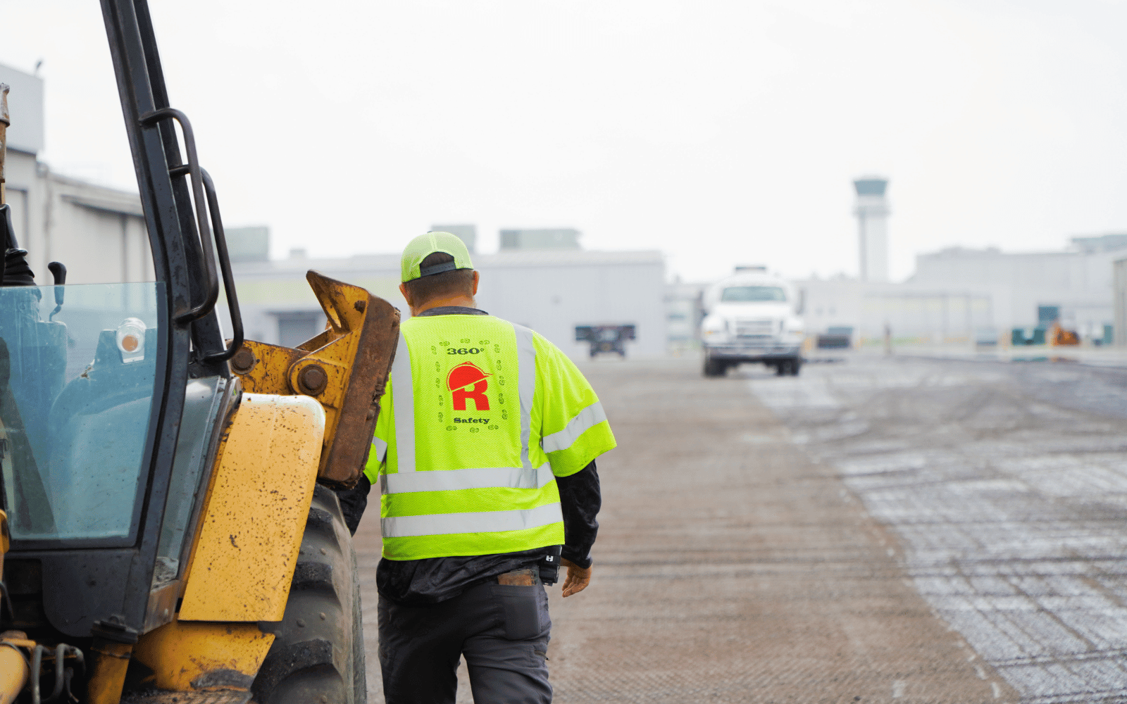 A construction worker in a neon yellow safety vest and cap stands next to heavy machinery on a worksite, with buildings and a white truck visible in the background.