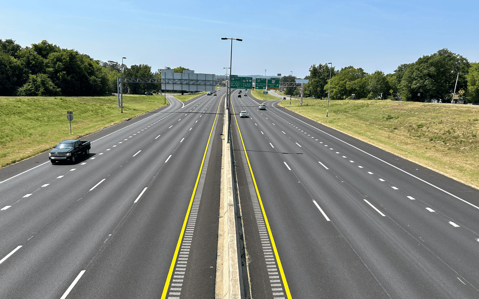 A wide view of an empty highway with six lanes in each direction, divided by a concrete barrier, bordered by grassy areas and trees under a clear blue sky. Only a few cars are visible on the road.