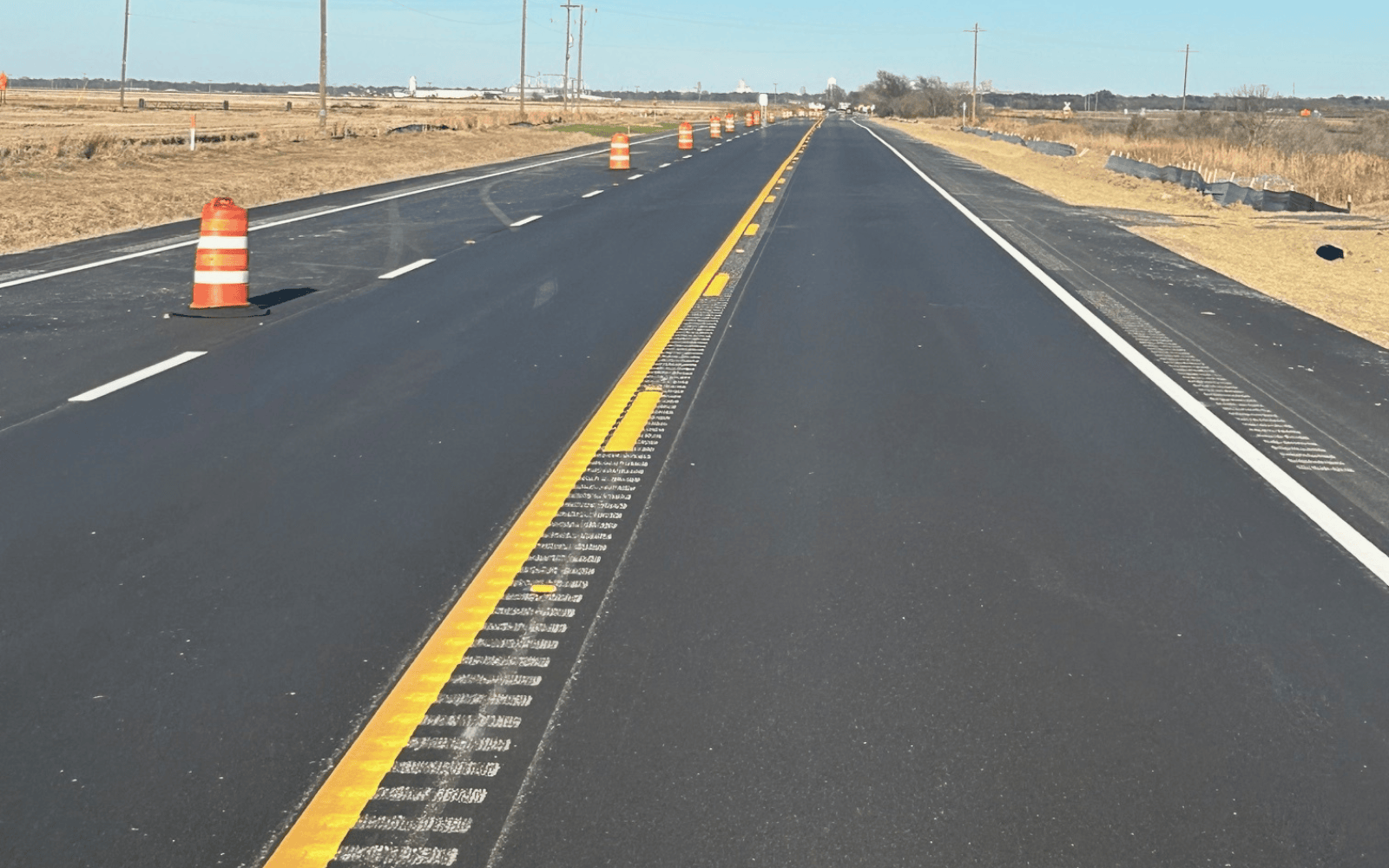 A newly paved, empty road stretches into the distance, marked with yellow and white lines. Orange construction barrels line the road, and fields with dry grass are visible on both sides under a clear sky.