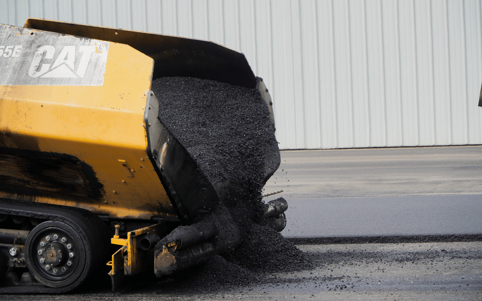 24 A yellow asphalt paver machine pours fresh black asphalt onto a road surface in front of a large, light gray metal building.