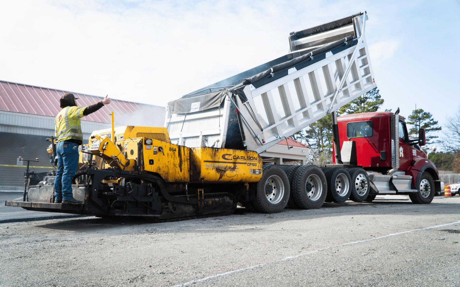 6 A construction worker operates a yellow asphalt paver next to a red dump truck with its bed raised, unloading material onto a road at a construction site.