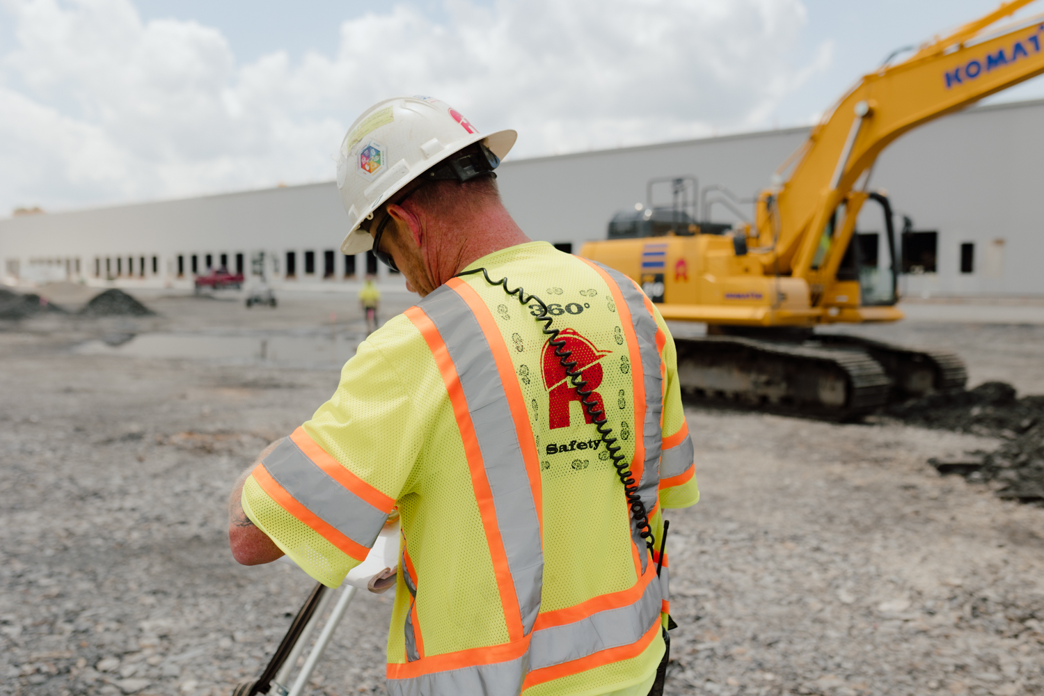 A construction worker in a safety vest and helmet operates surveying equipment at an outdoor worksite, with an excavator and a large building in the background.