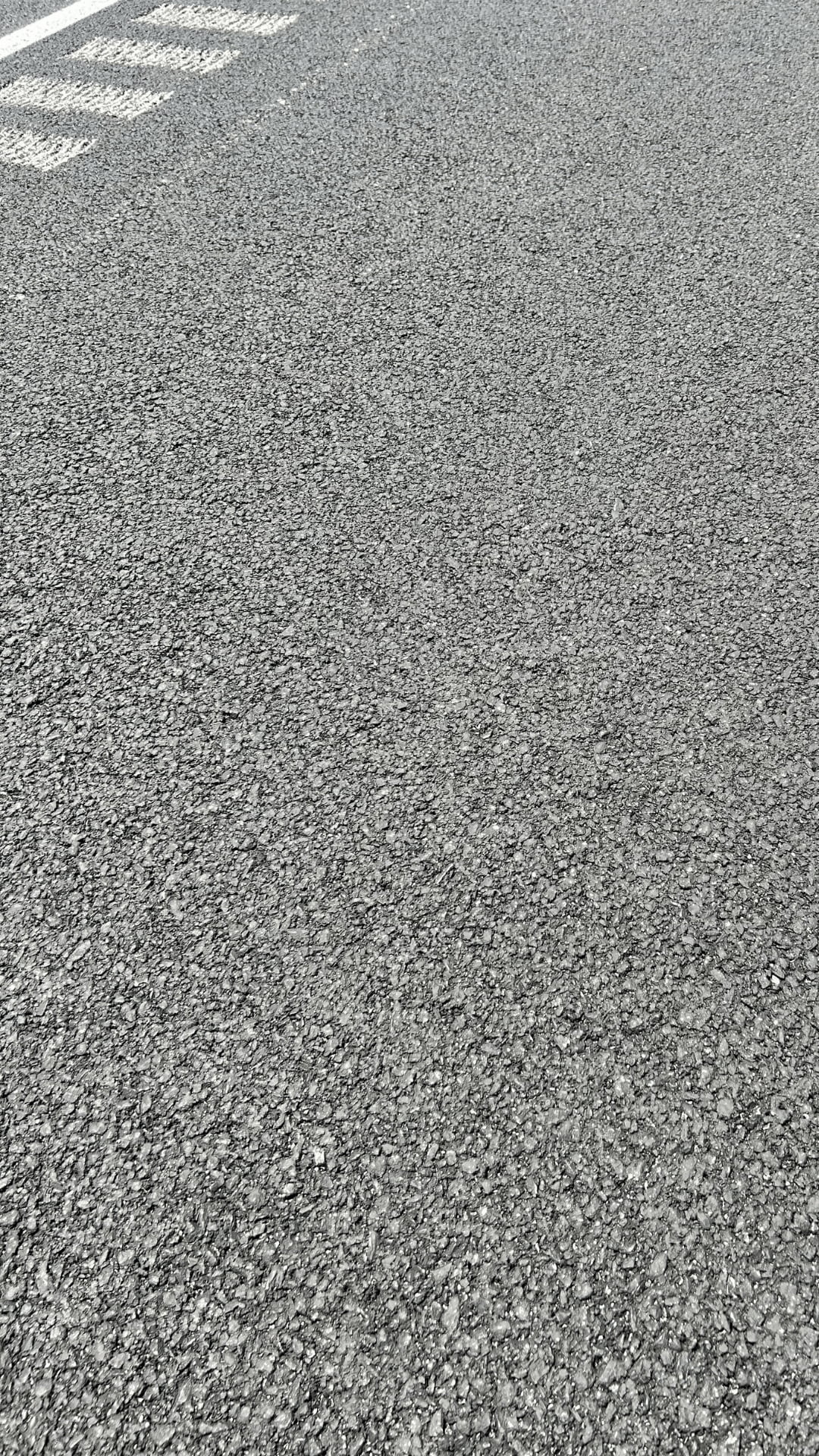 Close-up view of a paved road with visible asphalt texture, a white painted line, and partial white road markings near the top left corner.