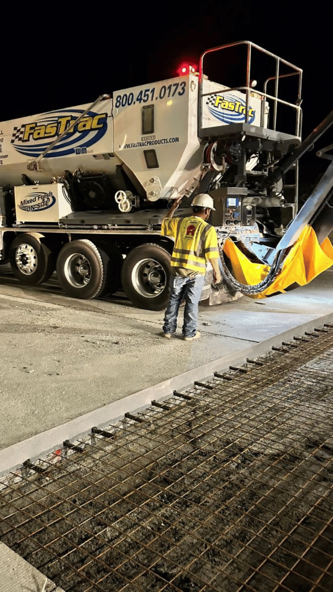 A construction worker in a yellow safety vest and helmet observes concrete being poured from a Fastrac cement truck onto a rebar grid at a nighttime work site.