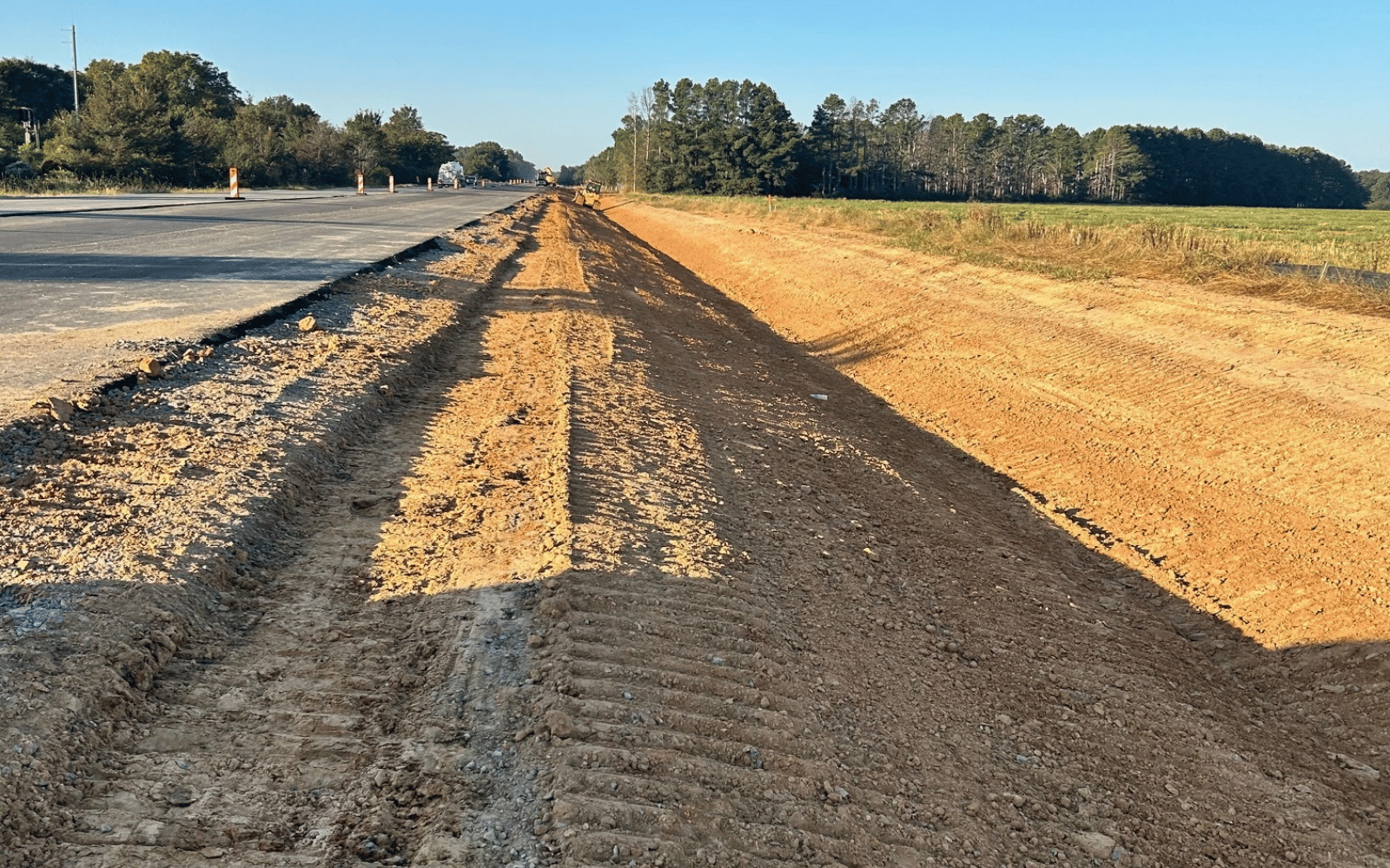 A road construction site with a newly graded dirt path beside a partially paved road, bordered by trees and open land under a clear blue sky.