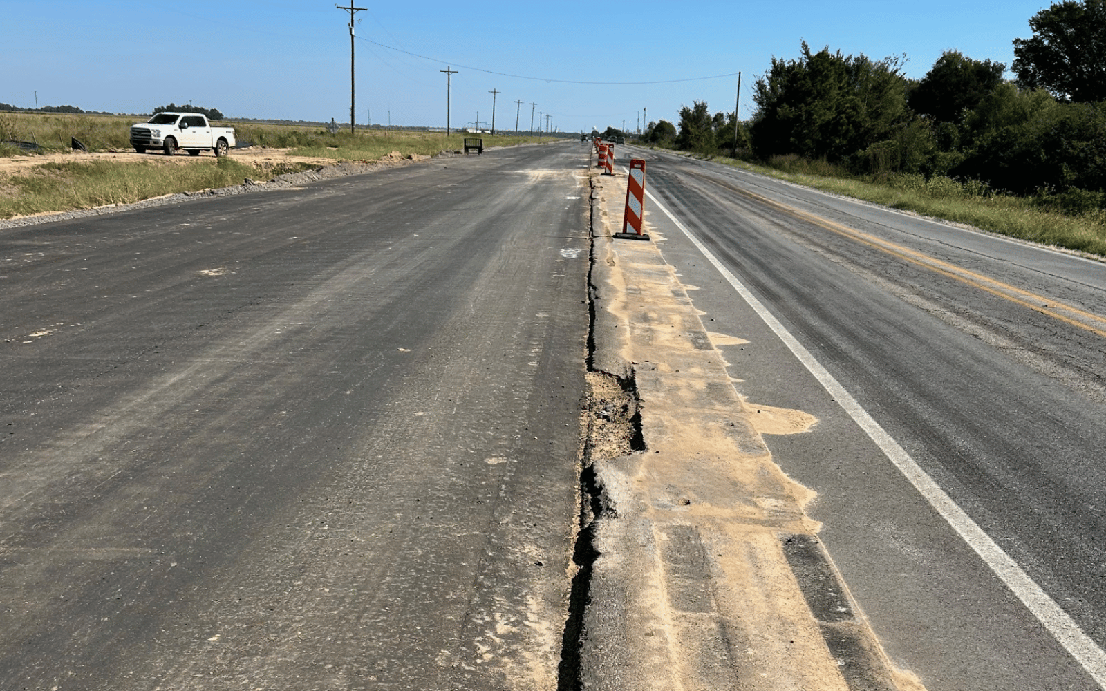 A rural road under construction with a rough, uneven surface and a raised dirt strip. Traffic cones and a construction sign mark the area. A white pickup truck is parked off to the side under a clear blue sky.