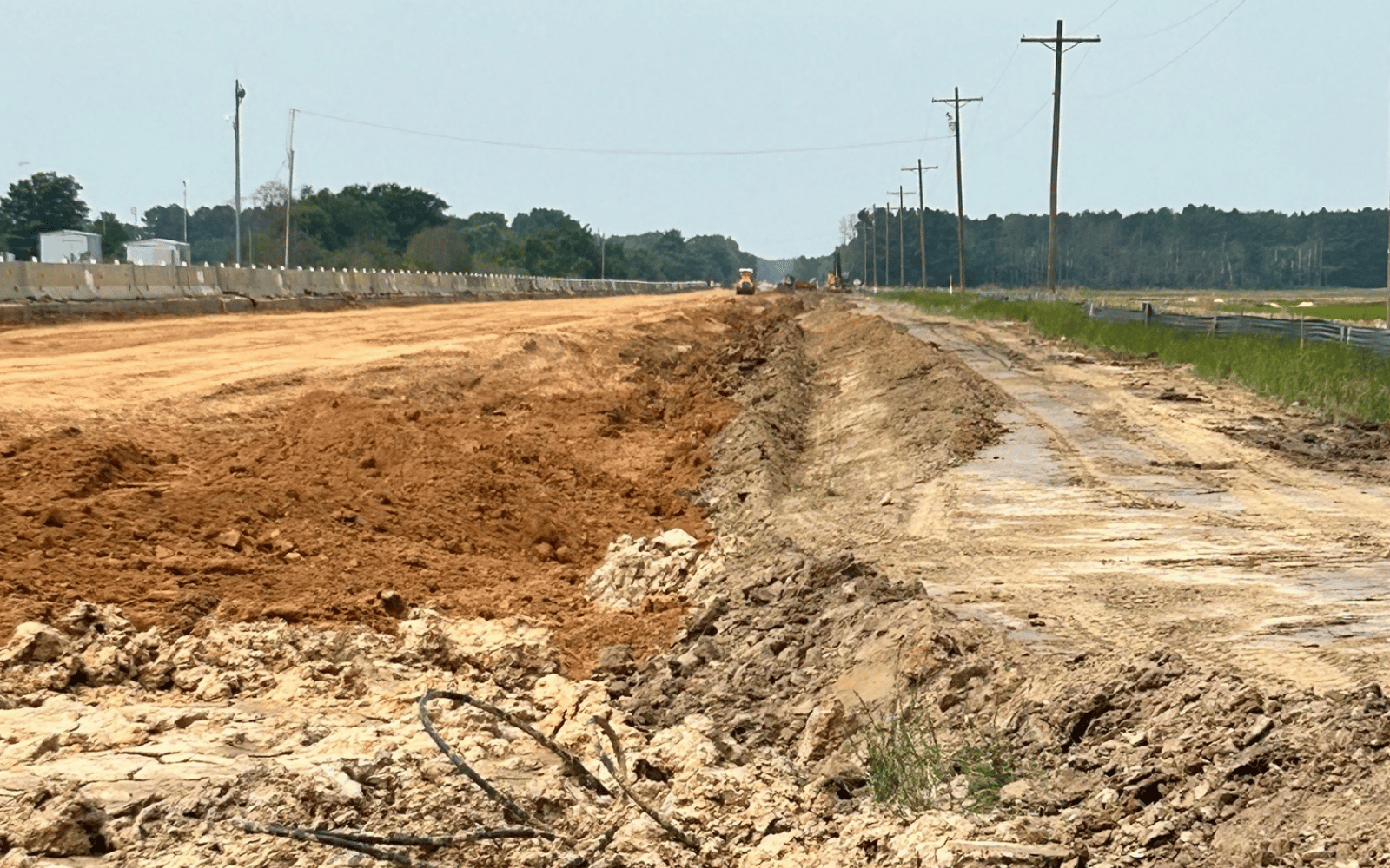 A wide, muddy construction site with dirt mounds and heavy machinery in the distance, running parallel to power lines and a rural road under a clear sky.