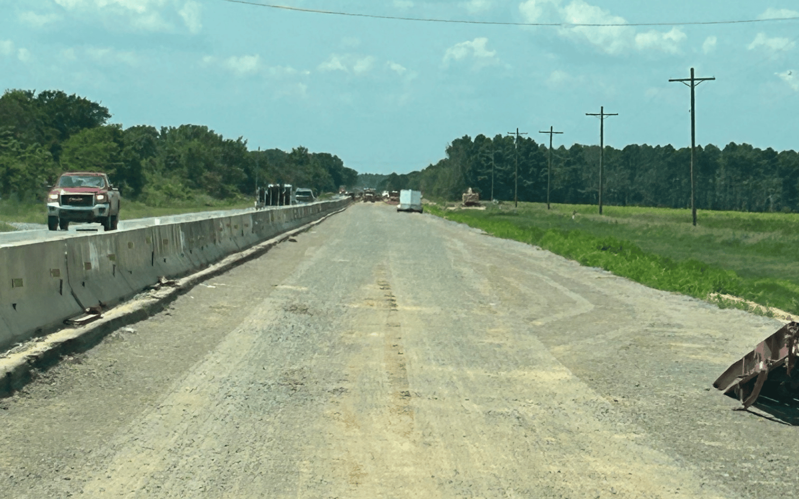 A gravel road under construction runs parallel to a highway with traffic barriers on the left and grassy fields and utility poles to the right, under a blue sky with scattered clouds.