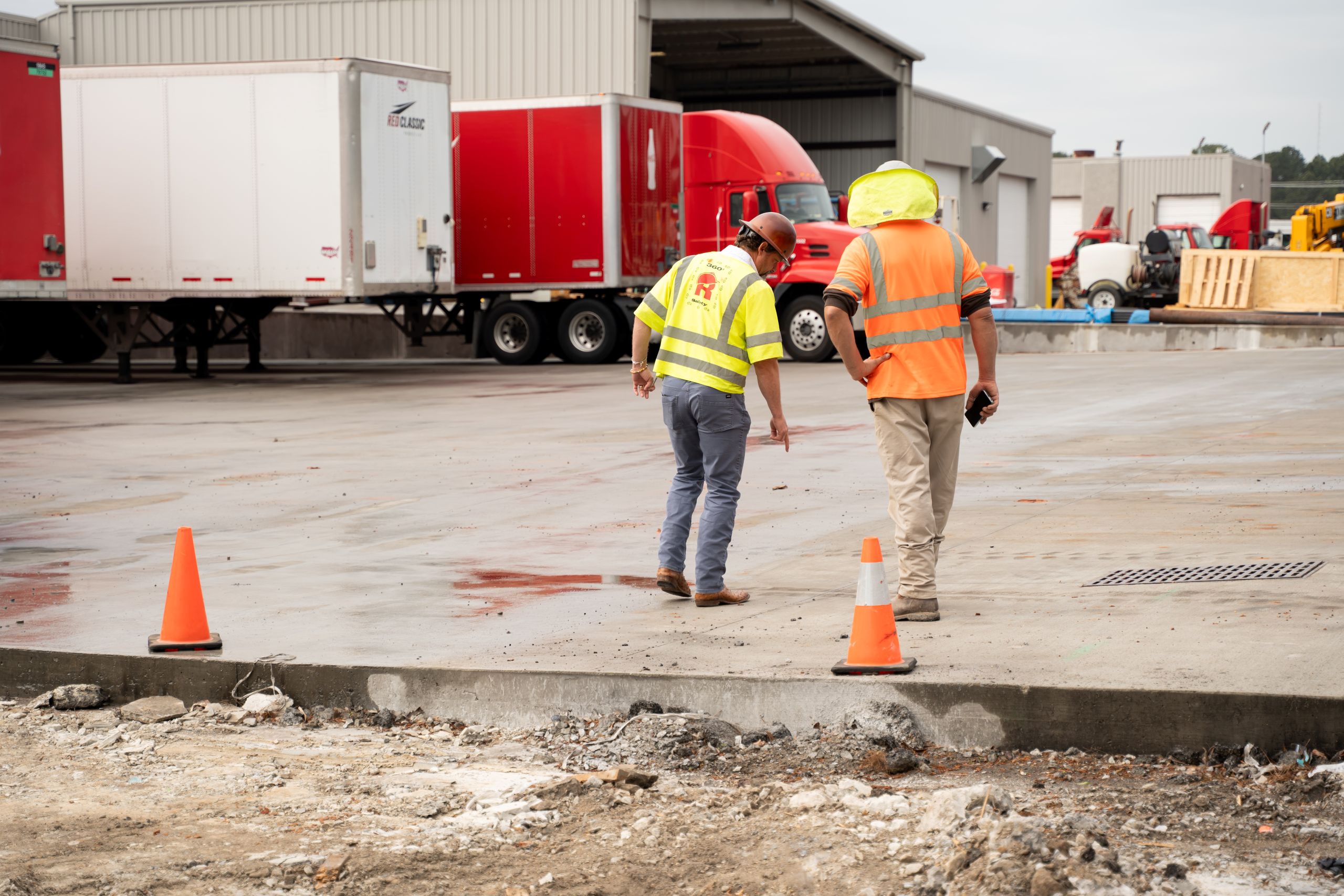 Two construction workers in safety vests and hard hats stand on a concrete surface near orange cones, discussing cement vs concrete while inspecting the ground at an industrial site with red trucks and buildings in the background.