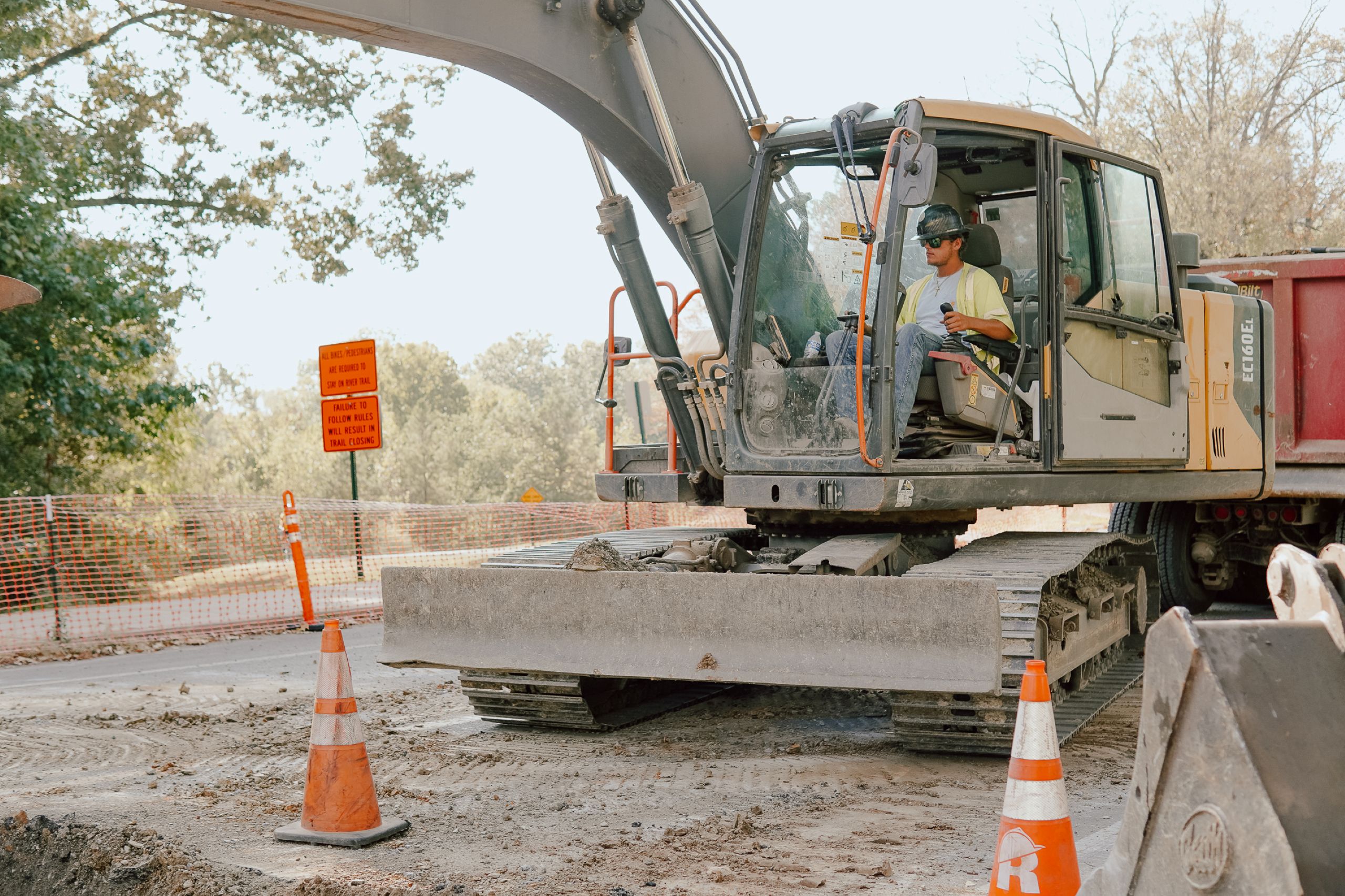 A construction worker operates an excavator on a road site, surrounded by orange traffic cones and caution signs, illustrating how roads are made. Trees and construction fencing are visible in the background.