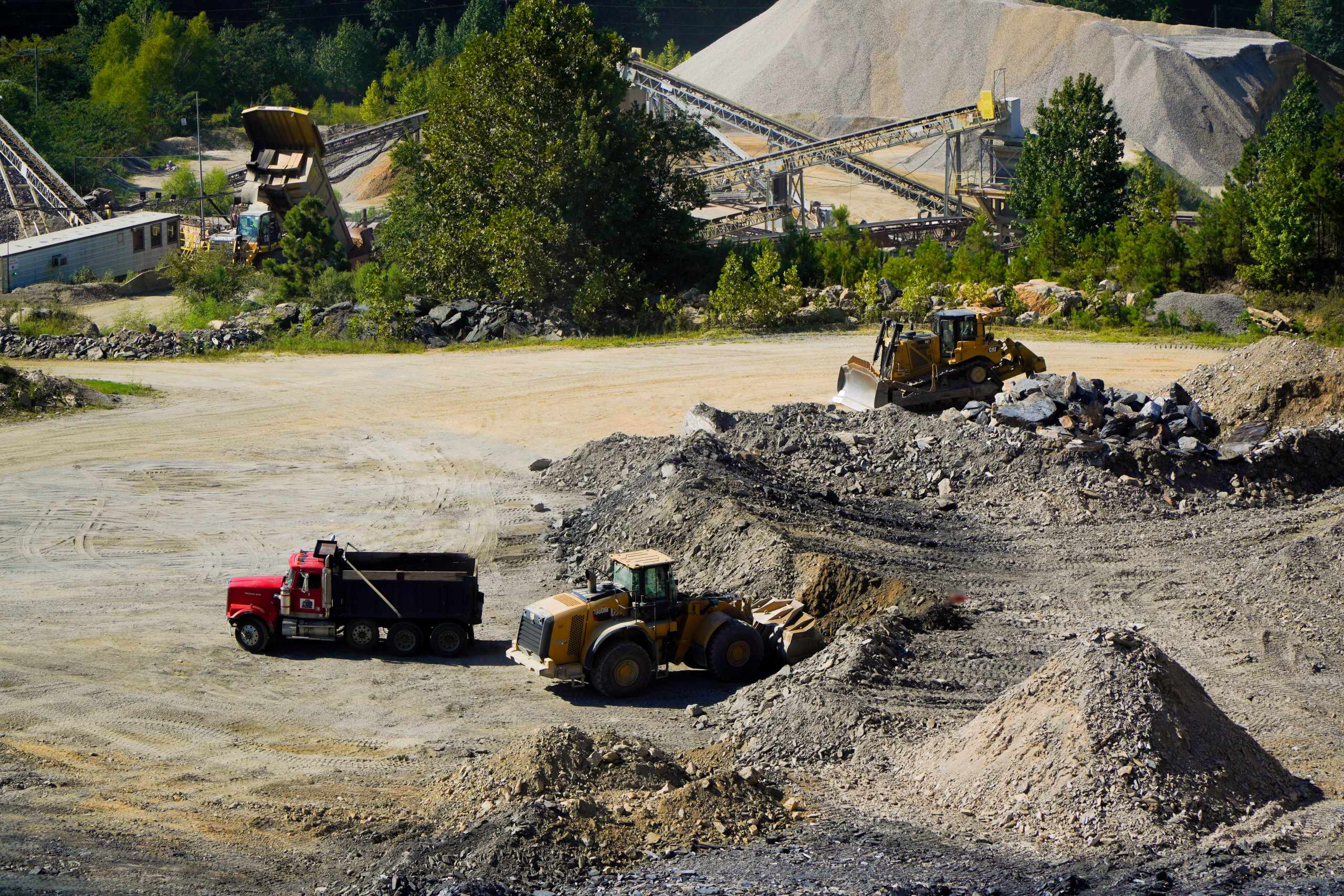 A dump truck and two bulldozers work at a quarry with piles of gravel, dirt, trees, and conveyor belts visible in the background under sunny weather, ensuring the structural integrity of the site is maintained throughout the operation.