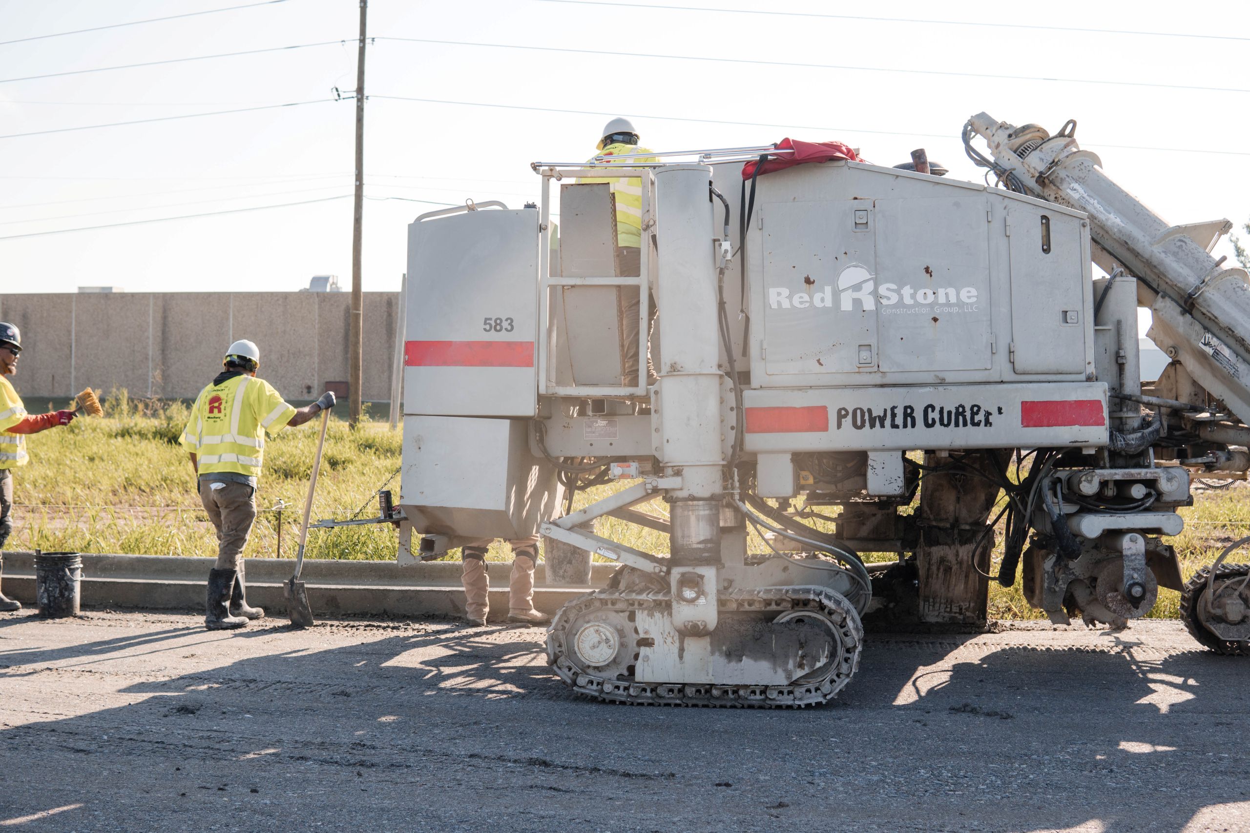 Construction workers operate a large gray road paving machine labeled Red Stone and Power Curber outdoors on a sunny day, with some workers using shovels near the curb.