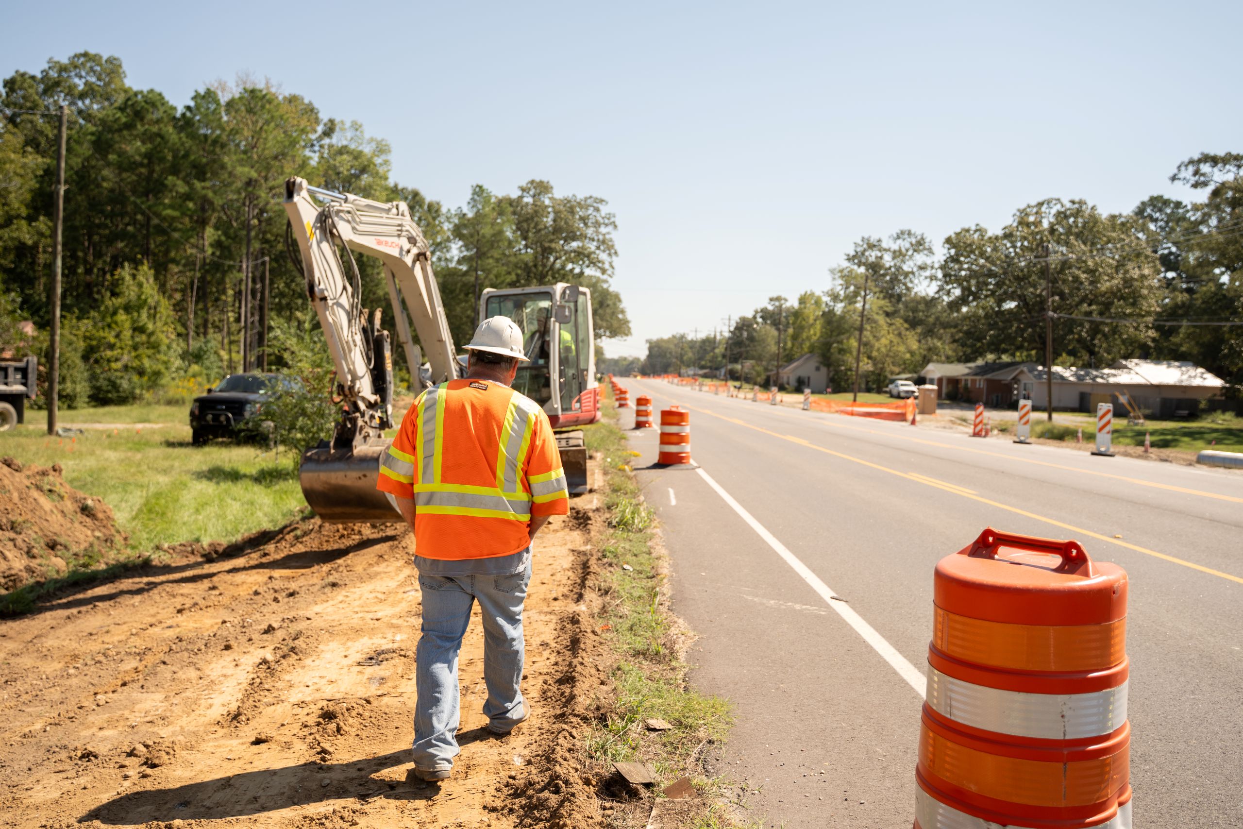 A construction worker in a safety vest and helmet walks near an excavator on a roadside. Orange traffic barrels line the road, and trees and houses are visible in the background under a clear sky.