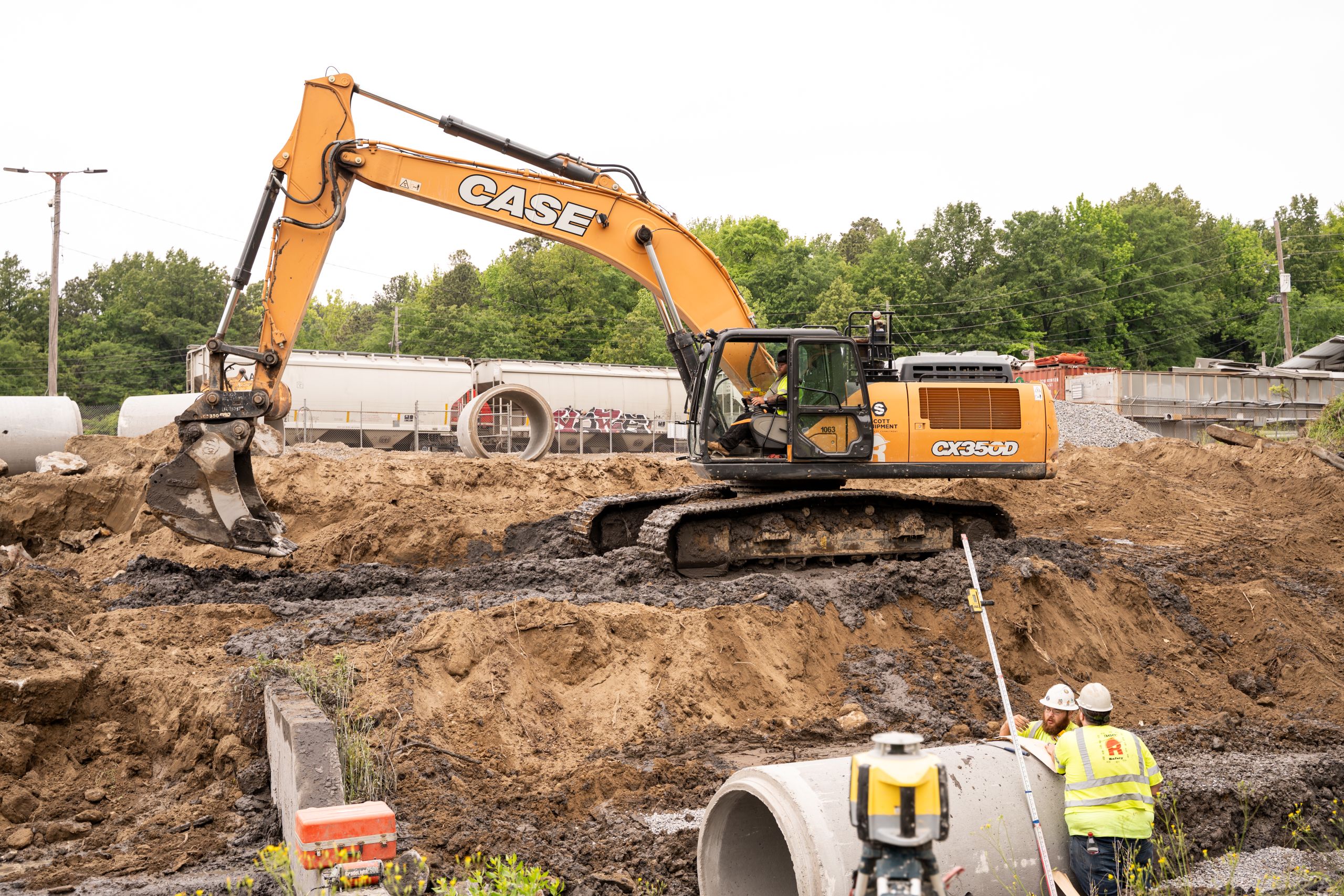 A Case CX350D excavator sits on a dirt pile at a construction site while two workers in high-visibility vests stand near concrete pipes.